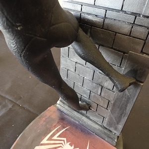 K&ouml;nnte beinhalten: Schwarze Figur eines Superhelden, der eine Backsteinmauer hochklettert. Die Figur hat strukturierte Oberfl&auml;chen und befindet sich &uuml;ber einem Sockel mit einem wei&szlig;en Spinnenemblem auf rotem und violettem Hintergrund.
