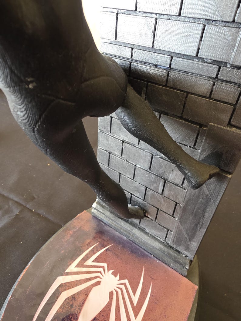 K&ouml;nnte beinhalten: Schwarze Figur eines Superhelden, der eine Backsteinmauer hochklettert. Die Figur hat strukturierte Oberfl&auml;chen und befindet sich &uuml;ber einem Sockel mit einem wei&szlig;en Spinnenemblem auf rotem und violettem Hintergrund.
