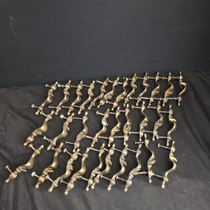 May include: A collection of twenty-four brass-toned metal drawer pulls, each featuring a curved, twisted design with a central ribbed detail and two attached mounting screws extending from the back of the hardware pieces.