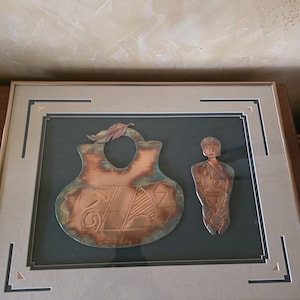 May include: Framed copper art piece featuring two figures: a stylized vase with leaf detail and a human-like form. The copper has a patina with hints of blue and green, set against a dark green background within a framed display.