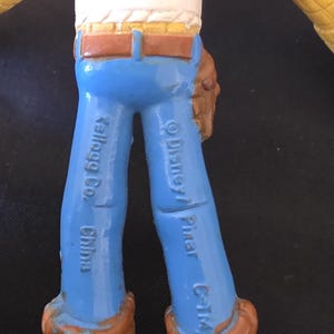 May include: A close-up of a Woody action figure from the Toy Story franchise. The figure has blue pants, brown boots, and a brown belt. The text "Kellogg Co. China" and "&copy; Disney/ Pixar C-T/M" are visible on the legs.