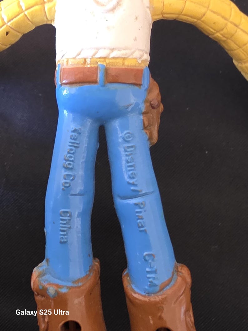 May include: A close-up of a Woody action figure from the Toy Story franchise. The figure has blue pants, brown boots, and a brown belt. The text "Kellogg Co. China" and "&copy; Disney/ Pixar C-T/M" are visible on the legs.