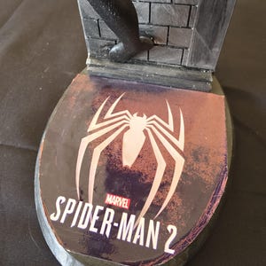 K&ouml;nnte beinhalten: Schwarze und graue Spider-Man 2-Statuenbasis mit dem Marvel-Logo und einem wei&szlig;en Spinnensymbol. Die Basis hat ein Backsteinmauer-Design und eine schwarze Figur, die die Wand hochklettert.