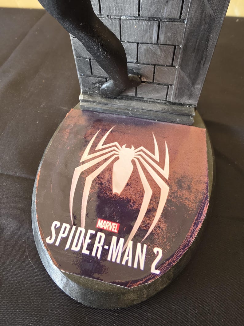 K&ouml;nnte beinhalten: Schwarze und graue Spider-Man 2-Statuenbasis mit dem Marvel-Logo und einem wei&szlig;en Spinnensymbol. Die Basis hat ein Backsteinmauer-Design und eine schwarze Figur, die die Wand hochklettert.