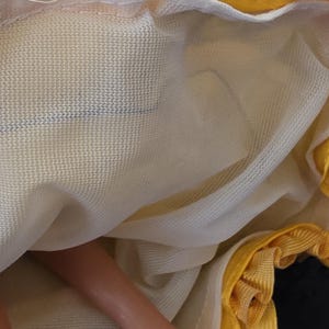 以下が含まれることがあります： 人形の黄色と白のドレスのクローズアップ。ドレスはフリル付きの黄色のトリムと白いメッシュのオーバーレイが付いています。人形の足が見え、生地は質感があるように見えます。