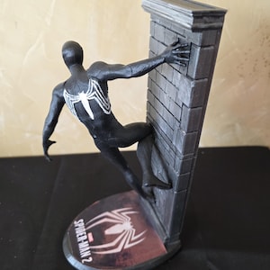 K&ouml;nnte beinhalten: Eine schwarze Spider-Man 2-Figur in dynamischer Pose, die sich an einer grauen Backsteinmauer festh&auml;lt. Die Figur hat ein wei&szlig;es Spinnenemblem auf dem R&uuml;cken. Der Sockel zeigt das Spider-Man 2-Logo und ein Spinnennetz-Design.