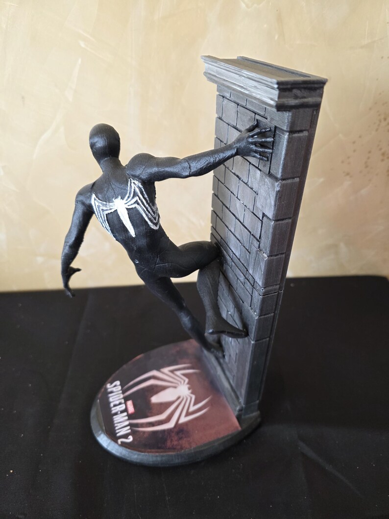 K&ouml;nnte beinhalten: Eine schwarze Spider-Man 2-Figur in dynamischer Pose, die sich an einer grauen Backsteinmauer festh&auml;lt. Die Figur hat ein wei&szlig;es Spinnenemblem auf dem R&uuml;cken. Der Sockel zeigt das Spider-Man 2-Logo und ein Spinnennetz-Design.