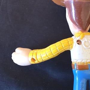 May include: A close-up of a Woody action figure from the Toy Story franchise. The figure has a brown cowboy hat, a yellow shirt with a white vest, blue jeans, and a tan face. The arm is extended, and the figure is made of plastic.