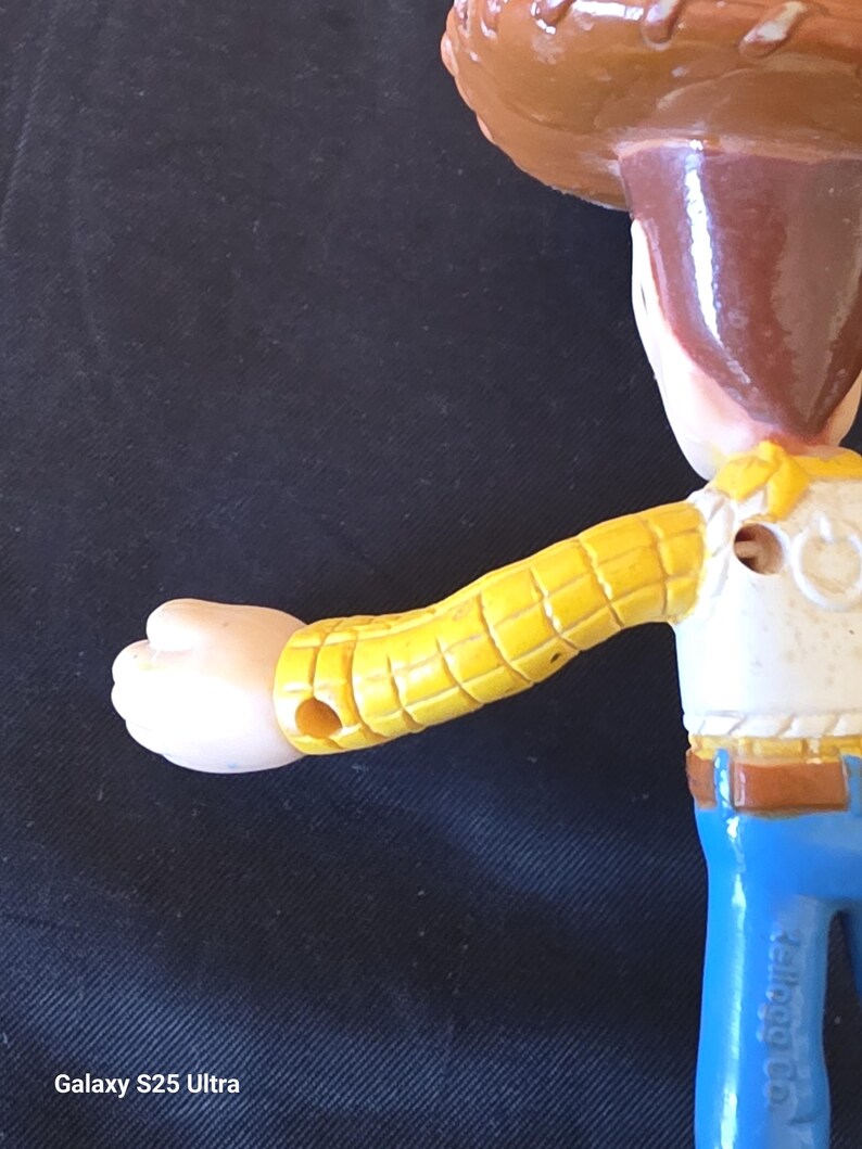 May include: A close-up of a Woody action figure from the Toy Story franchise. The figure has a brown cowboy hat, a yellow shirt with a white vest, blue jeans, and a tan face. The arm is extended, and the figure is made of plastic.