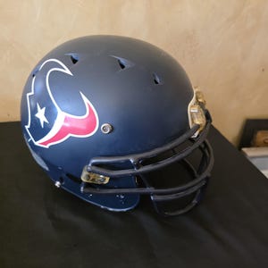 Puede incluir: Un casco de fútbol americano azul marino oscuro con el logotipo de los Houston Texans en el lateral, que consiste en una cabeza de toro estilizada en azul marino, rojo y blanco. El casco incluye una máscara facial azul marino y herrajes metálicos.
