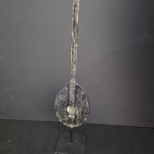 May include: A decorative metal sculpture of a lute with a distressed black and white finish. The instrument features a long neck, a rounded body with a central circular sound hole, and is mounted vertically on a square black metal base stand.