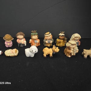 May include: A collection of twelve small, hand-painted resin figurines depicting a nativity scene. The set includes figures of people in robes, a baby in a manger, a white sheep, a brown cow, and a donkey, all featuring soft, muted colors.