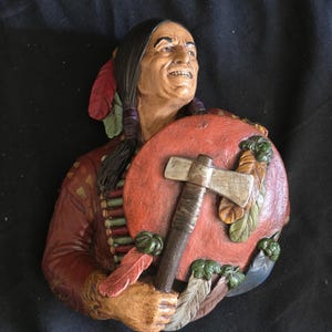 May include: A three-dimensional wall sculpture depicting a person with long dark hair, wearing a red garment with fringe details. The figure holds a round red shield adorned with feathers and a silver-toned tomahawk with a wooden handle.