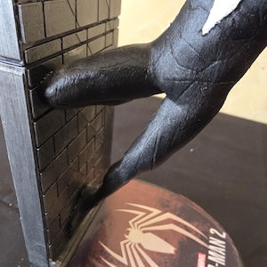 K&ouml;nnte beinhalten: Eine schwarz-wei&szlig;e Spider-Man 2-Statue mit einer Figur in dynamischer Pose, mit einem strukturierten schwarzen Anzug und wei&szlig;en Akzenten. Die Figur interagiert mit einer Backsteinmauer. Der Sockel zeigt das Spider-Man 2-Logo.
