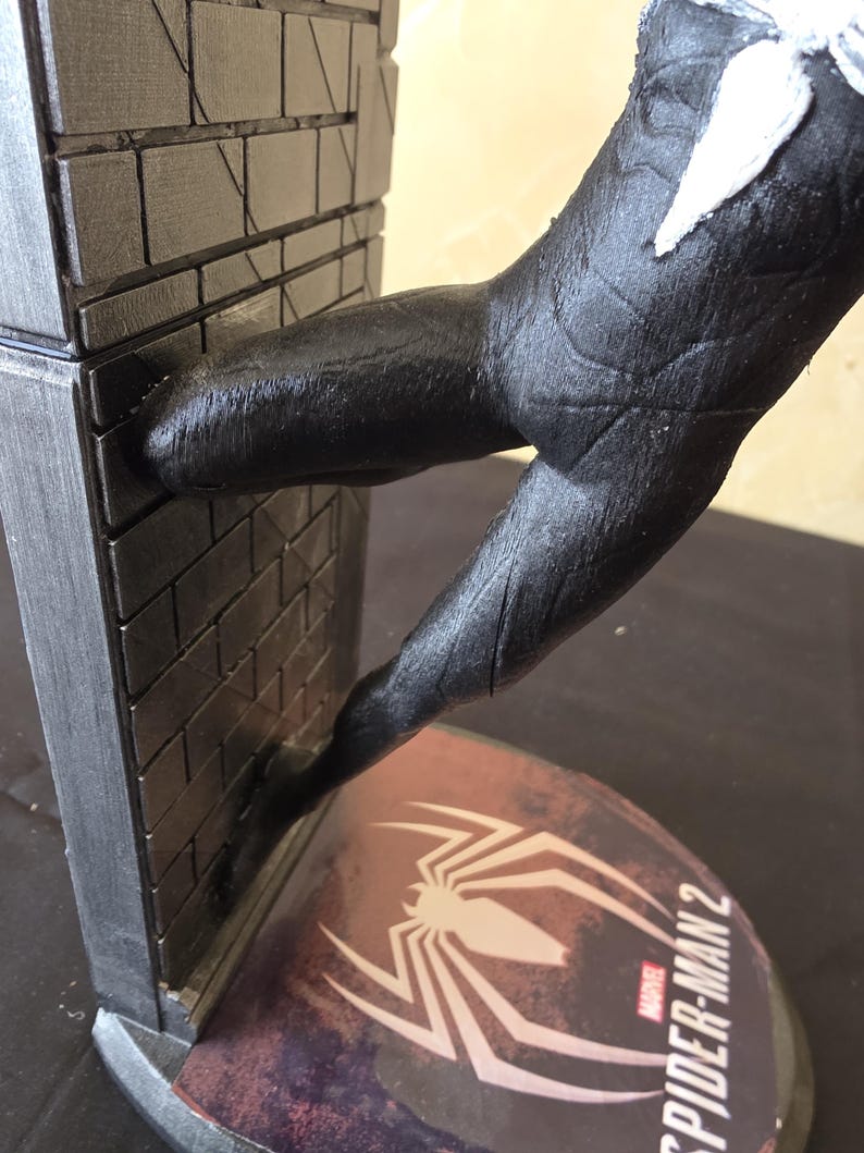 K&ouml;nnte beinhalten: Eine schwarz-wei&szlig;e Spider-Man 2-Statue mit einer Figur in dynamischer Pose, mit einem strukturierten schwarzen Anzug und wei&szlig;en Akzenten. Die Figur interagiert mit einer Backsteinmauer. Der Sockel zeigt das Spider-Man 2-Logo.
