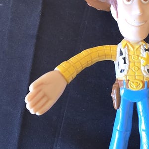 May include: A Woody action figure, featuring a brown cowboy hat, yellow shirt with black and white accents, blue jeans, and brown boots. The figure has a sheriff's badge and a holster. The figure is a popular character from the Toy Story franchise.