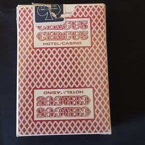 Puede incluir: Una baraja de cartas rectangular en una caja blanca con un patrón de rombos rojos. La caja presenta el texto Circus Circus Hotel/Casino en letras rojas y un sello azul de la U.S. Playing Card Company en el borde superior.