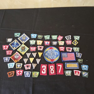 Puede incluir: Una colección de parches bordados, incluyendo un gran parche circular "GREAT ACTIVITIES PATCH" y un parche con la bandera de EE. UU. Otros parches presentan varios diseños, incluyendo un lobo, un oso y el número "387". Los parches son de varias formas y colores.