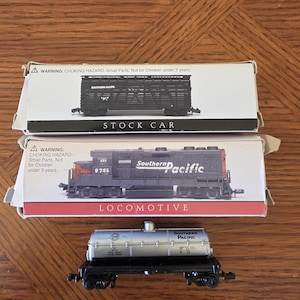 May include: Three model train cars on a wooden surface. The top car is a black stock car in a white box. Below is a black and red locomotive in a white box. The bottom car is a silver tank car.