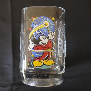 May include: Clear glass tumbler featuring Mickey Mouse in a sorcerer's outfit, holding a wand with a star. The design includes a blue background with fireworks and stars. The year "2000" is printed on the base. The Disney logo is also visible.