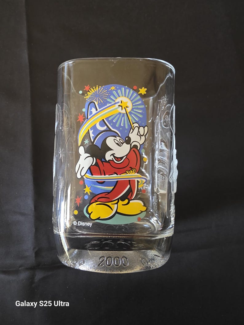May include: Clear glass tumbler featuring Mickey Mouse in a sorcerer's outfit, holding a wand with a star. The design includes a blue background with fireworks and stars. The year "2000" is printed on the base. The Disney logo is also visible.