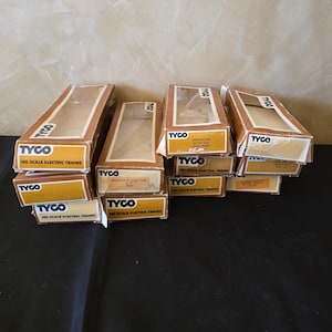 May include: A collection of ten rectangular cardboard boxes for Tyco HO scale electric trains. Each box features a clear plastic window on the top, brown and white branding, and printed labels identifying specific train car models like flat cars.