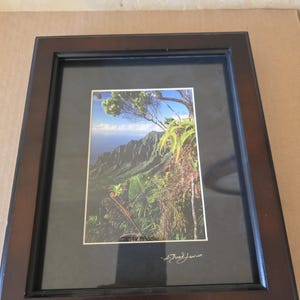 May include: Framed landscape print featuring a lush green mountain range under a blue sky. The print is set within a dark brown wooden frame with a black matting. Signed in the lower right corner.