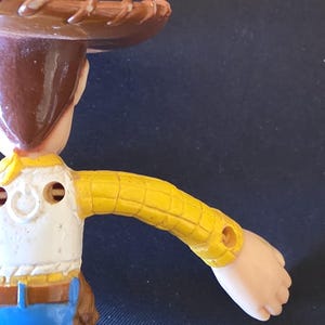 May include: A close-up of a Woody action figure from the Toy Story franchise. The figure is wearing a brown cowboy hat, a yellow plaid shirt, a white vest, and blue jeans. The figure's arm is extended. The text "Galaxy S25 Ultra" is visible.