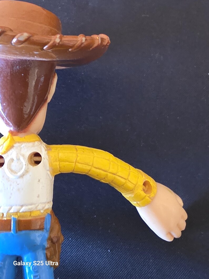 May include: A close-up of a Woody action figure from the Toy Story franchise. The figure is wearing a brown cowboy hat, a yellow plaid shirt, a white vest, and blue jeans. The figure's arm is extended. The text "Galaxy S25 Ultra" is visible.