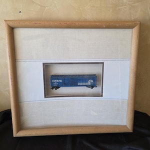 May include: Framed miniature blue Conrail train car model. The model is displayed in a shadow box with a white background and a light brown wooden frame. The text "CONRAIL" is visible on the side of the train car.