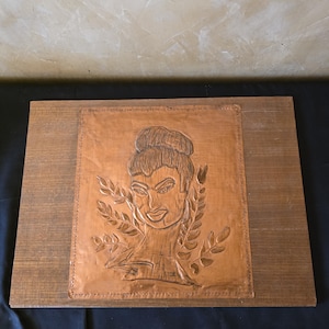 May include: A rectangular wooden plaque featuring a centered, embossed copper sheet. The copper panel displays a hand-tooled portrait of a person with an updo hairstyle, surrounded by stylized leaf motifs, with a decorative dotted border.