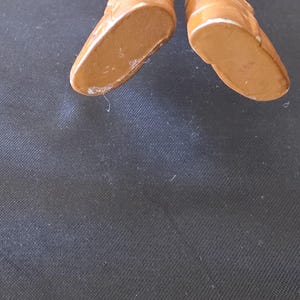May include: Close-up of two brown ceramic doll shoes with white trim. The shoes have a glossy finish and are set against a dark background. The soles of the shoes are visible.