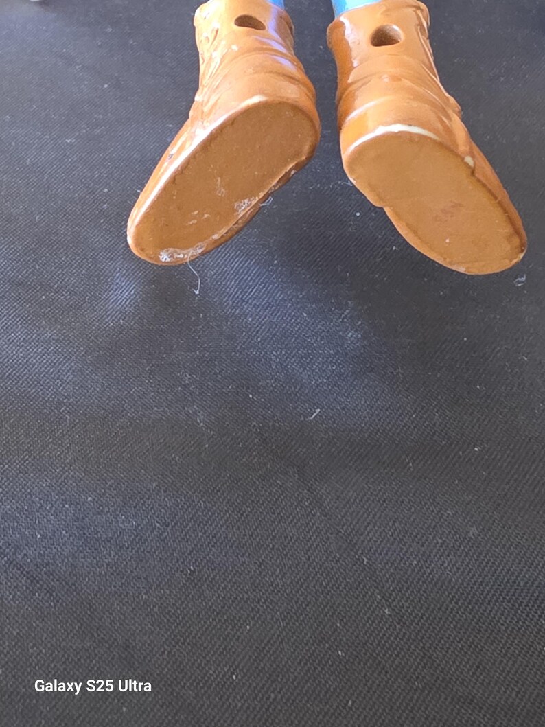 May include: Close-up of two brown ceramic doll shoes with white trim. The shoes have a glossy finish and are set against a dark background. The soles of the shoes are visible.