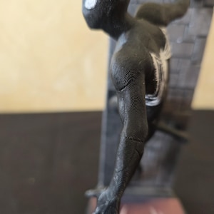 K&ouml;nnte beinhalten: Eine schwarze Spider-Man-Figur, die an einer strukturierten Wand haftet. Die Figur hat wei&szlig;e Augendetails und ein wei&szlig;es Spinnenemblem auf der Brust. Der Sockel zeigt ein Spider-Man-Logo. Die Figur ist ein 3D-gedrucktes Objekt.