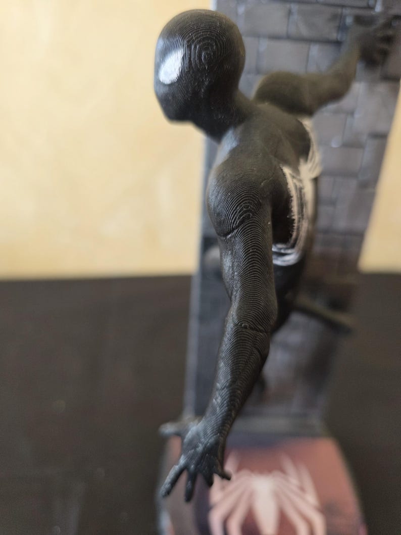 K&ouml;nnte beinhalten: Eine schwarze Spider-Man-Figur, die an einer strukturierten Wand haftet. Die Figur hat wei&szlig;e Augendetails und ein wei&szlig;es Spinnenemblem auf der Brust. Der Sockel zeigt ein Spider-Man-Logo. Die Figur ist ein 3D-gedrucktes Objekt.
