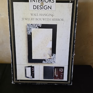 May include: A rectangular product box for an Interiors by Design wall hanging jewelry box with mirror. The box features a black frame with white floral accents, displaying the product closed and open with interior storage hooks and shelves.