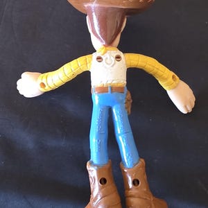 May include: A Woody action figure from the Toy Story franchise. The figure is wearing a brown cowboy hat, a yellow plaid shirt, a white vest, blue jeans, and brown boots. The figure has its arms outstretched.