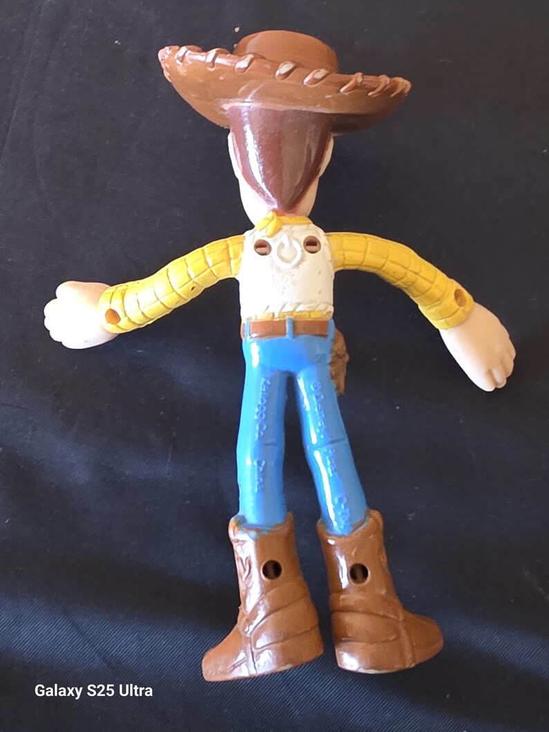 May include: A Woody action figure from the Toy Story franchise. The figure is wearing a brown cowboy hat, a yellow plaid shirt, a white vest, blue jeans, and brown boots. The figure has its arms outstretched.