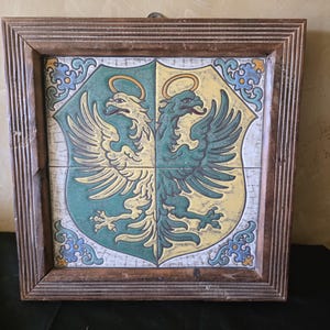 May include: A framed decorative tile featuring a heraldic design. The artwork is divided into two halves, one green and one yellow, each displaying a stylized eagle with a halo. The frame is made of dark brown wood.