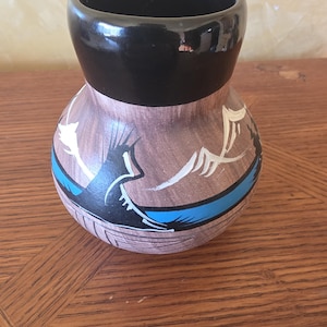 Puede incluir: Un jarrón de cerámica pintado a mano con borde negro y cuerpo marrón. El jarrón presenta una franja azul y diseños geométricos blancos y negros. El jarrón mide aproximadamente 13 cm de alto.