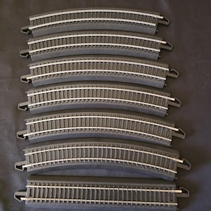 May include: Seven pieces of model train track with dark gray plastic roadbeds and silver metal rails. Six pieces are curved, and one piece is straight. Each track section features integrated plastic connectors at both ends for assembly.