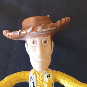 May include: A plastic Woody action figure from the Toy Story franchise. The figure has a brown cowboy hat, a yellow shirt, a vest with a sheriff's badge, and a smiling face. The text "Galaxy S25 Ultra" is visible.