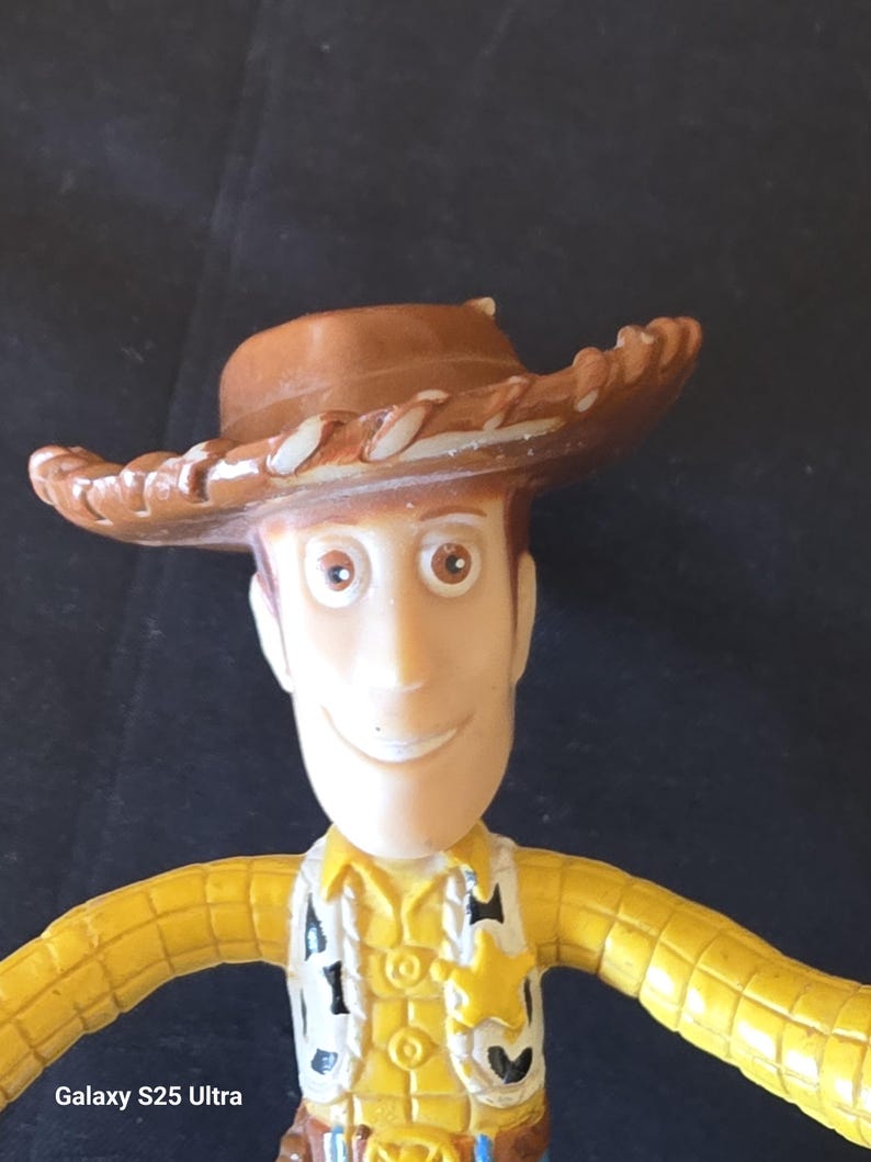 May include: A plastic Woody action figure from the Toy Story franchise. The figure has a brown cowboy hat, a yellow shirt, a vest with a sheriff's badge, and a smiling face. The text "Galaxy S25 Ultra" is visible.