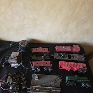 May include: A collection of vintage toy train components, including red and green train cars, a locomotive, track pieces, and electrical transformers. The items are arranged on a black surface, showcasing the various parts of a model train set.