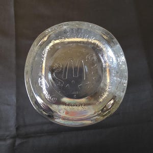 May include: Clear glass McDonald's drinking glass base with the McDonald's logo and text. The glass has a circular base with embossed details. The glass is on a black surface.