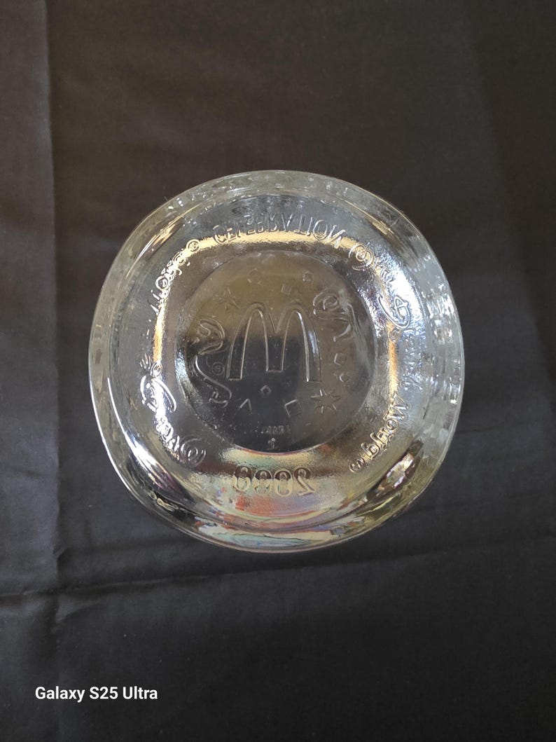May include: Clear glass McDonald's drinking glass base with the McDonald's logo and text. The glass has a circular base with embossed details. The glass is on a black surface.