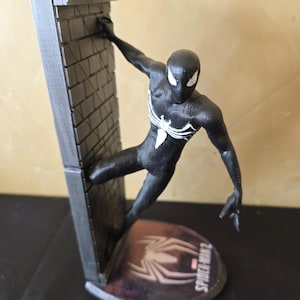 K&ouml;nnte beinhalten: Schwarze Spider-Man-Figur, die an einer grauen Backsteinmauer h&auml;ngt. Die Figur hat wei&szlig;e Augen und ein wei&szlig;es Spinnenemblem. Die Basis zeigt das Spider-Man 2-Logo.