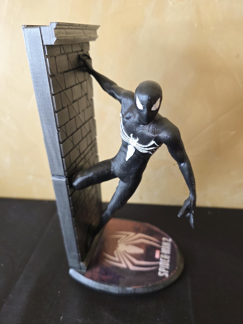K&ouml;nnte beinhalten: Schwarze Spider-Man-Figur, die an einer grauen Backsteinmauer h&auml;ngt. Die Figur hat wei&szlig;e Augen und ein wei&szlig;es Spinnenemblem. Die Basis zeigt das Spider-Man 2-Logo.