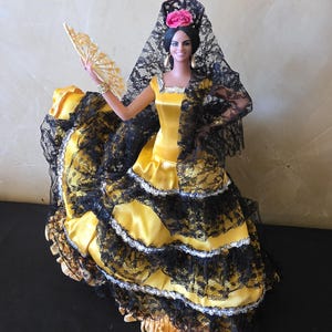 以下が含まれることがあります： 伝統的なスペインのフラメンコ衣装を着た人形。人形は、黒いレースのトリムが付いた黄色のサテンのドレス、黒いレースのショール、髪にピンクの花を身に着けています。彼女は扇子を持っています。