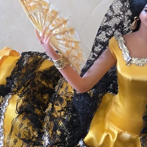 以下が含まれることがあります： 黒髪にピンクの花、金の装飾を施した、黒いレースのトリムが付いた黄色のドレスを着た人形。ドレスは首と袖に銀色のトリムが付いています。人形は扇子を持っており、扇子は黄色と白です。